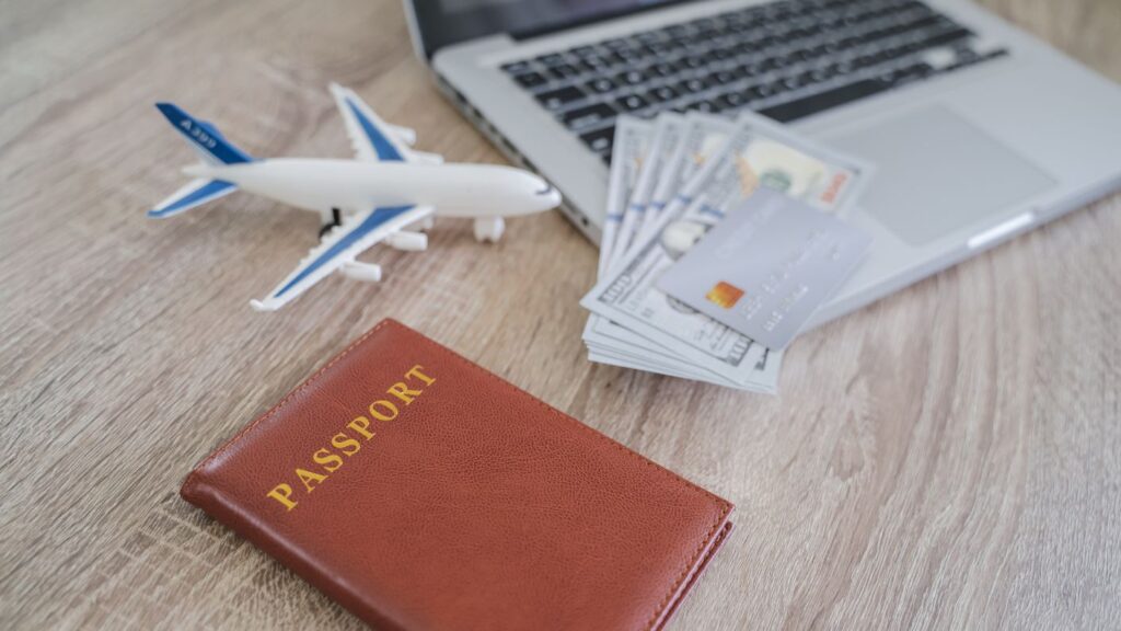 Foto di copertina con laptop, modellino di aereo, passaporto, carta di credito e banconote, simbolo di pianificazione di viaggi e prenotazioni online.