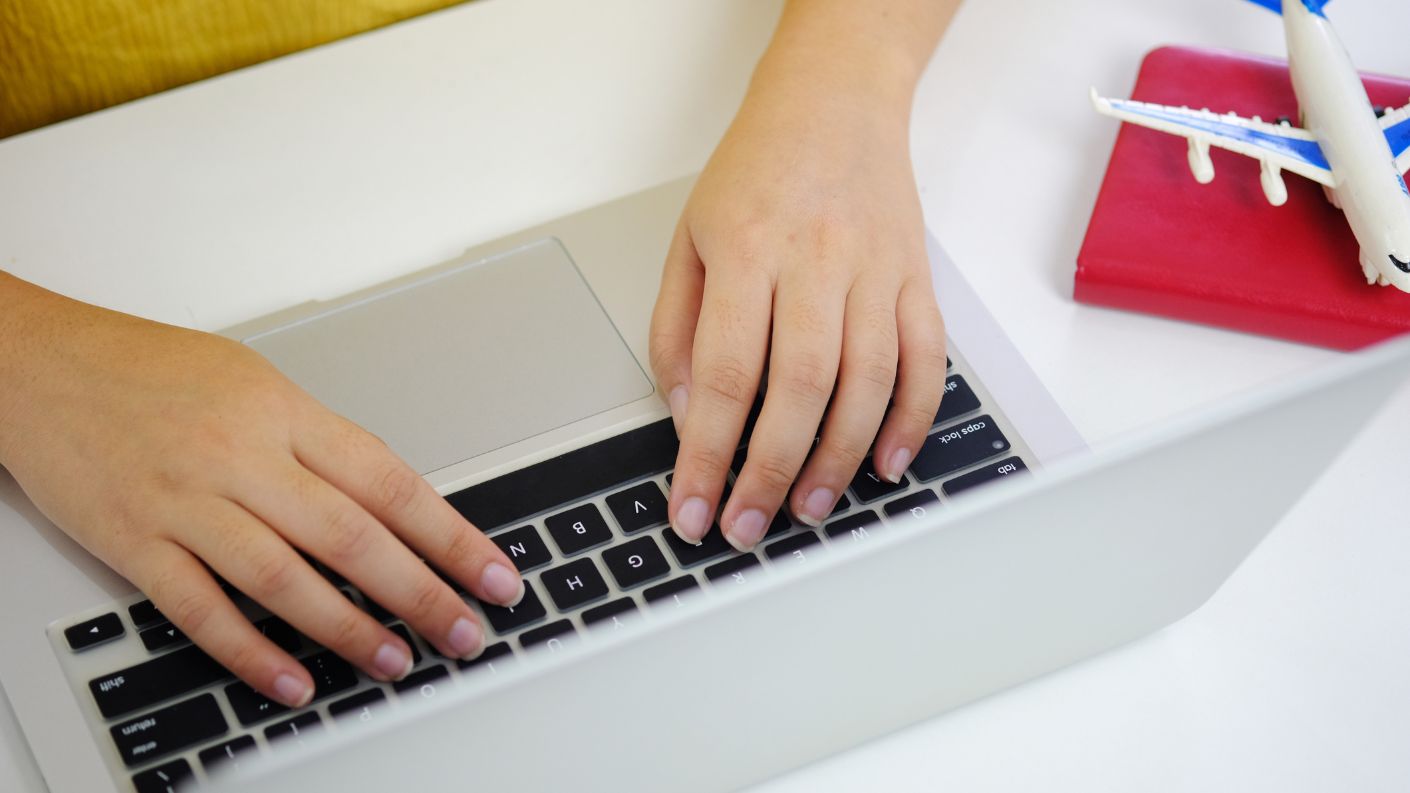 Mani che digitano sulla tastiera di un computer, accanto a un modellino di aereo e un passaporto, simbolo di prenotazioni di viaggi online.