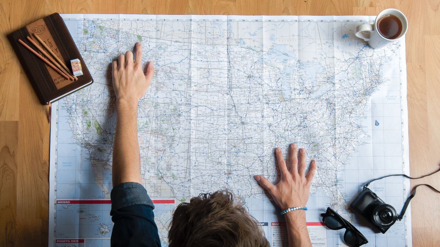 Uomo che indica una destinazione su una mappa, con occhiali da sole, tazza di caffè e quaderno con matite sul tavolo, simbolo di pianificazione di viaggi e avventure.