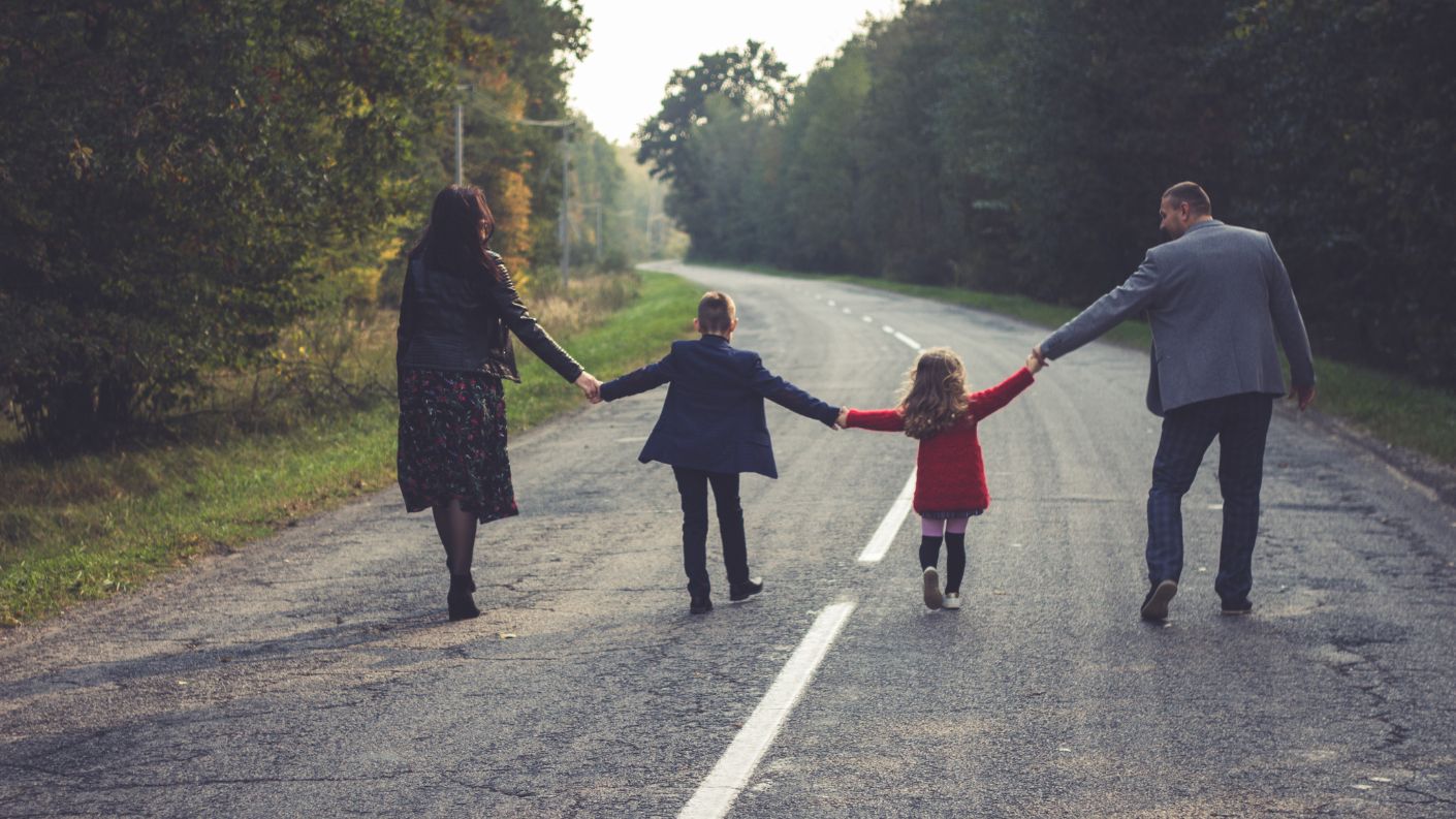 Famiglia in mezzo alla strada che si tiene per mano, con i due bambini al centro, simbolo di viaggio e unione familiare.
