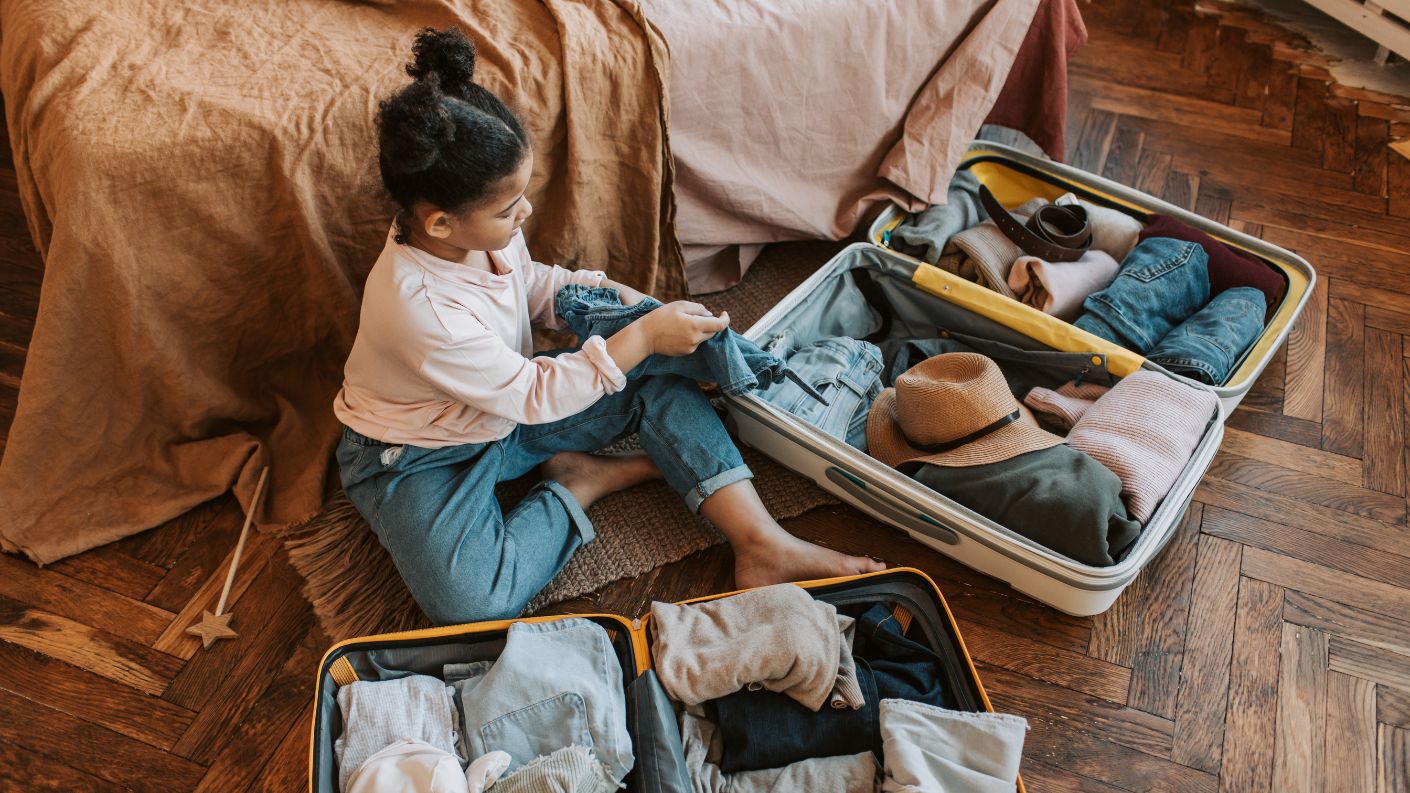 Bambina seduta sul pavimento che prepara una valigia aperta, accanto un'altra valigia pronta per il viaggio, simbolo di preparativi e avventure in famiglia.