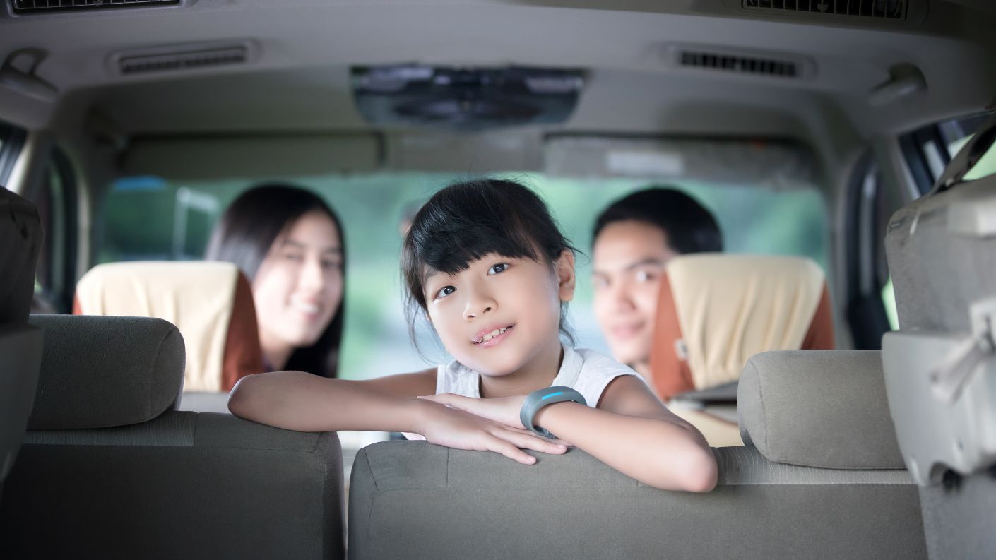 Bambina sorridente che guarda verso l'obiettivo dall'interno di un'auto, con i genitori sorridenti sui sedili anteriori, simbolo di viaggio in famiglia e felicità.